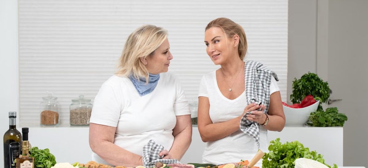 Two women in a kitchen preparing fresh vegetables and cheese with olive oil and spices.